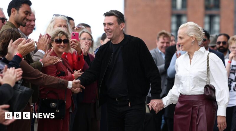 British Prime Minister Andy Burnham dressed in all black and shaking hands with a supporter in a crowed, while holding his other hand with his wife Marie-France van Heel, who is dressed in a white shirt and burgundy leather skirt.
