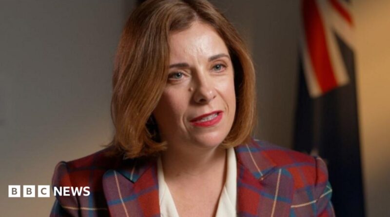 A woman in a tartan jacket and white dress speaks. Several flags can be seen in the background
