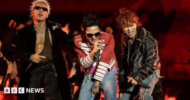 Three performers stand at the front of a stage holding handheld microphones during a live performance. One performer wears a long black coat and sunglasses, another wears a red military-style jacket with white trim and jeans, and a third wears a black studded leather jacket and ripped jeans. The stage is lit in red, with flames or flame-effect visuals displayed behind the performers.