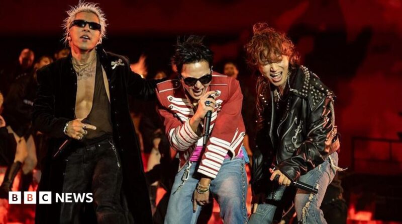 Three performers stand at the front of a stage holding handheld microphones during a live performance. One performer wears a long black coat and sunglasses, another wears a red military-style jacket with white trim and jeans, and a third wears a black studded leather jacket and ripped jeans. The stage is lit in red, with flames or flame-effect visuals displayed behind the performers.