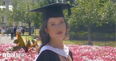 A girl with curly dark hair, wearing a white dress and black graduation cap and gown. She is stood in front of a flowerbed of pink flowers, with trees and people in the background, and is smiling at the camera.