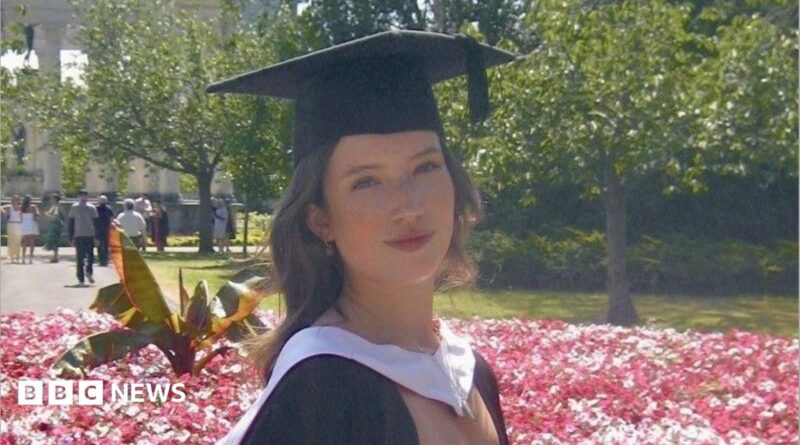 A girl with curly dark hair, wearing a white dress and black graduation cap and gown. She is stood in front of a flowerbed of pink flowers, with trees and people in the background, and is smiling at the camera.