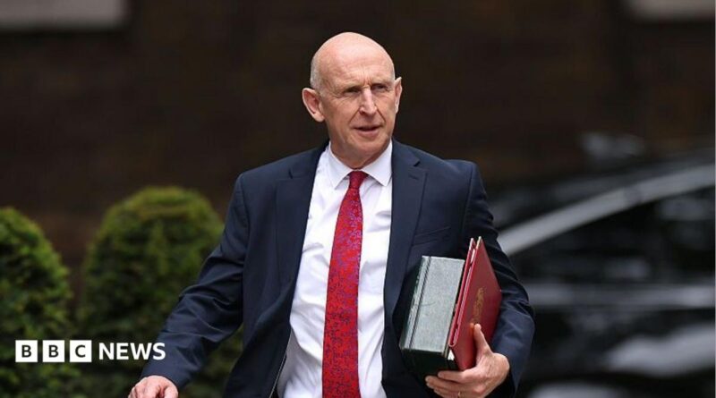 An image of John Healey from the waist up. He is walking and wearing a dark suit with a white shirt and red patterned tie. He is carrying a red folder. The background is blurred by you can see that he is outside from the outline of bushes.