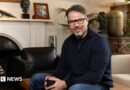 Chris Rokos sitting in a room wearing a dark blue jumper and blue jeans. In the background there is a fireplace, sofa and chest of drawers.