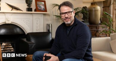 Chris Rokos sitting in a room wearing a dark blue jumper and blue jeans. In the background there is a fireplace, sofa and chest of drawers.