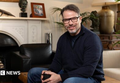 Chris Rokos sitting in a room wearing a dark blue jumper and blue jeans. In the background there is a fireplace, sofa and chest of drawers.