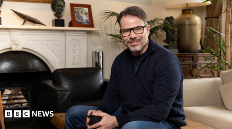 Chris Rokos sitting in a room wearing a dark blue jumper and blue jeans. In the background there is a fireplace, sofa and chest of drawers.