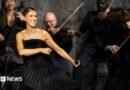 Dion dancing in a black dress, holding a microphone, while musicians play behind her