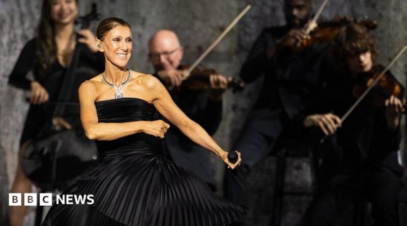 Dion dancing in a black dress, holding a microphone, while musicians play behind her