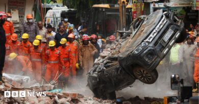 National Disaster Response Force (NDRF) personnel look on at a building collapse in Delhi, while a crushed car is pulled by a digger from the rubble.