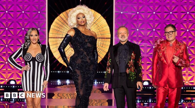 A promotional shot for Smoggie Queens which shows three men in drag posing in a conservatory alongside two women.  In the foreground, one person sits on a chair wearing a blonde wig, fishnet tights, a patterned blue top and black lace-up boots, while another sits on the floor in camouflage shorts, black boots and a burgundy vest with badges. Behind them, three people stand wearing a denim outfit, a purple dress, and a green sequined dress with bright feathered sleeves. The room contains rainbow curtains, pink feather boas, costume accessories, and a white mannequin head wearing bunny ears on a small table to the right.