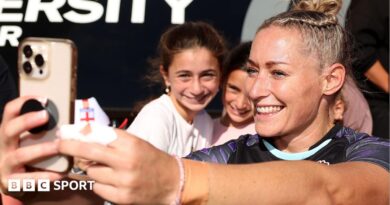 Natasha Hunt, wearing a blue England shirt, smiles while taking a selfie with a crowd of fans behind her