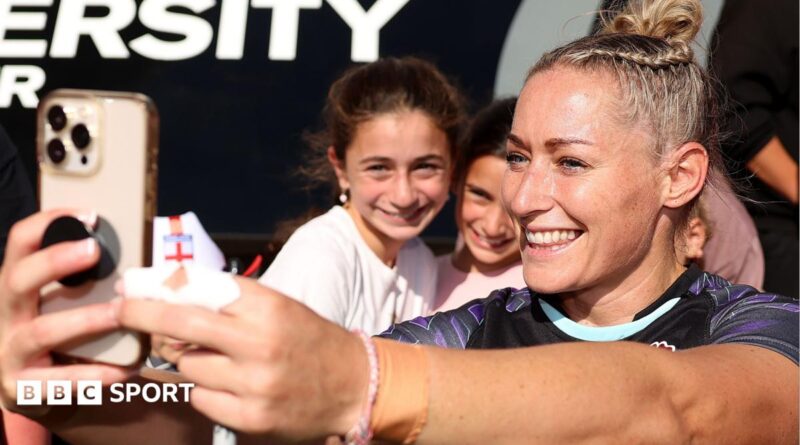 Natasha Hunt, wearing a blue England shirt, smiles while taking a selfie with a crowd of fans behind her