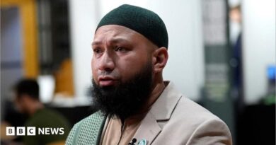 Mothin Ali looking to his right, wearing a beige suit jacket with a green and white scarf draped over his right shoulder