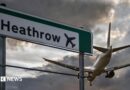 A commercial aeroplane flying low on final approach to land at Heathrow Airport, passing directly over an overhead highway road sign.