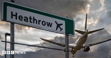 A commercial aeroplane flying low on final approach to land at Heathrow Airport, passing directly over an overhead highway road sign.