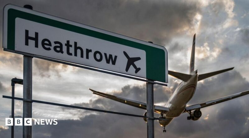 A commercial aeroplane flying low on final approach to land at Heathrow Airport, passing directly over an overhead highway road sign.