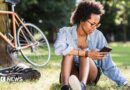 Student sits on the grass with a phone in her hand, a tablet computer on the grass and a bike resting on a tree behind her.