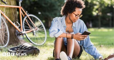 Student sits on the grass with a phone in her hand, a tablet computer on the grass and a bike resting on a tree behind her.