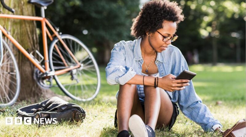 Student sits on the grass with a phone in her hand, a tablet computer on the grass and a bike resting on a tree behind her.