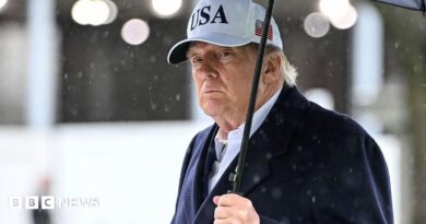 President Trump holds an umbrella as rain drops fall around him. He wears a dark suit and a white baseball cap with USA written on the front and an American flag on the side.