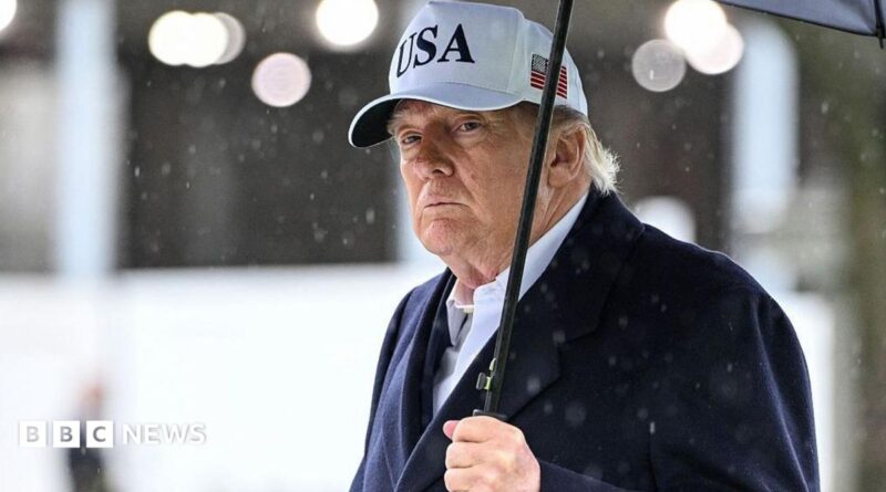 President Trump holds an umbrella as rain drops fall around him. He wears a dark suit and a white baseball cap with USA written on the front and an American flag on the side.