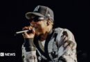 Jay-Z: Stars join US rapper at long-awaited UK show Jay-Z raps into a microphone while performing on stage. He wears a black and white cardigan and a New York Yankees hat.
