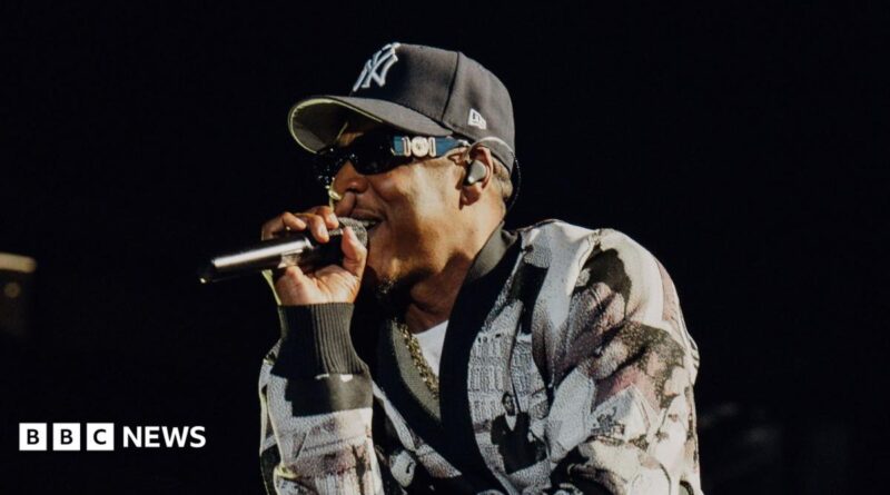 Jay-Z raps into a microphone while performing on stage. He wears a black and white cardigan and a New York Yankees hat.
