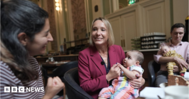 Helen Morgan is in the centre of the picture, smiling and holding a baby on her lap. Morgan has a blonde bob and is wearing a berry-coloured jacket over a navy top. She is looking at a woman in a stripy top on the left and a man holding another baby is on the right.