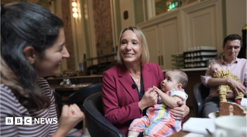 Helen Morgan is in the centre of the picture, smiling and holding a baby on her lap. Morgan has a blonde bob and is wearing a berry-coloured jacket over a navy top. She is looking at a woman in a stripy top on the left and a man holding another baby is on the right.