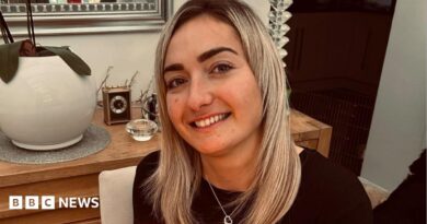 Maddy Cusack pictured smiling at home. She has long blonde hair worn loose and wears a silver heart necklace over a black top. She's pictured inside, in front of a side board with ornaments and plants