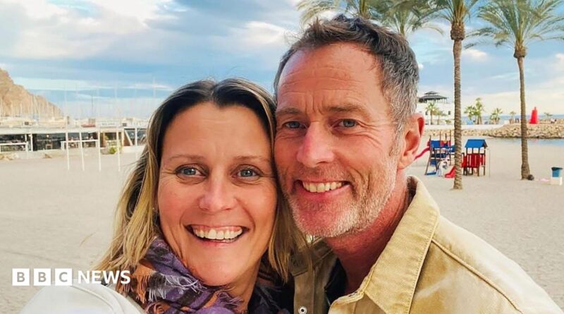A couple on a beach. It is sunny and the sand is white. Behind them are some trees.