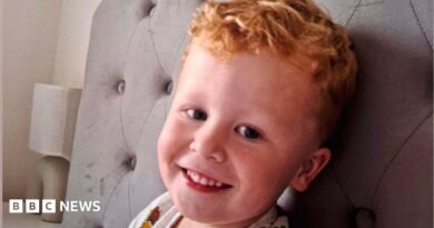 Three-year-old Noah Woods, with light hair and wearing a brightly coloured top, smiles at the camera.