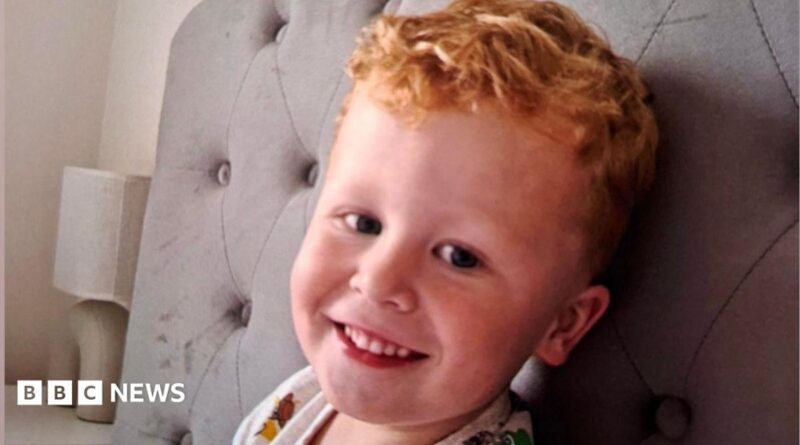 Three-year-old Noah Woods, with light hair and wearing a brightly coloured top, smiles at the camera.
