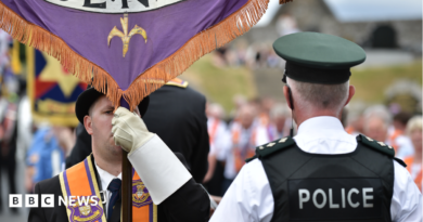 A man is wearing an orange sash and a black bowler hat and holding a banner. A policeman has his back to the camera. He has a black vest on stating police in white lettering. He has a cap and a white shirt on. A parade is out of focus in the background.