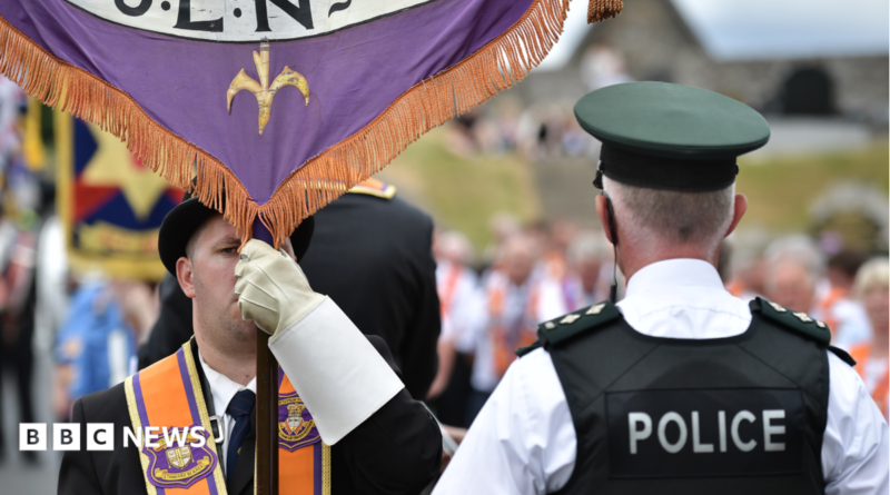 A man is wearing an orange sash and a black bowler hat and holding a banner. A policeman has his back to the camera. He has a black vest on stating police in white lettering. He has a cap and a white shirt on. A parade is out of focus in the background.
