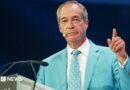 Nigel farage, wearing a light blue suit and white shirt, gestures with his left hand 
