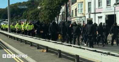 Dozens of men dressed in black and wearing face coverings, being escorted down a dual carriageway in Dover by uniformed police officers.