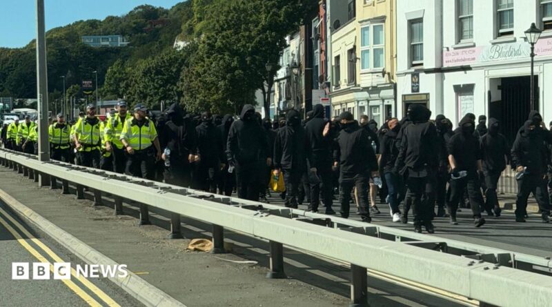 Dozens of men dressed in black and wearing face coverings, being escorted down a dual carriageway in Dover by uniformed police officers.