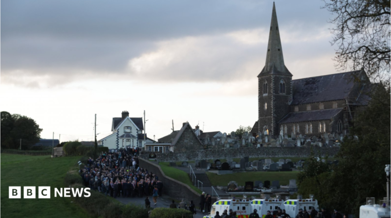 Orange Order members and their supporters gather near Drumcree Parish Church in Portadown. There are white police vehicles further down the road.