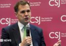 Robert Jenrick holding a microphone wearing a dark suit and a green tie. He is sitting in front of a branded sign for the Centre for Social Justice