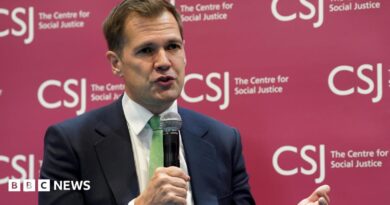 Robert Jenrick holding a microphone wearing a dark suit and a green tie. He is sitting in front of a branded sign for the Centre for Social Justice
