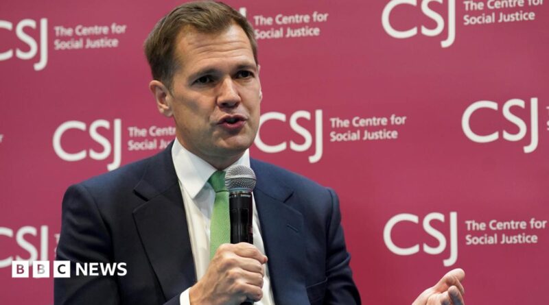 Robert Jenrick holding a microphone wearing a dark suit and a green tie. He is sitting in front of a branded sign for the Centre for Social Justice