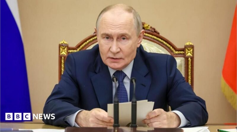 Putin sits in front of a desk and microphone holding some notes