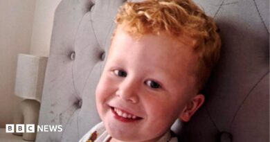 Three-year-old Noah Woods, with light hair and wearing a brightly coloured top, smiles at the camera.