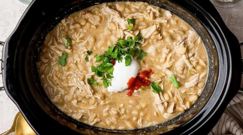 Slow cooker filled with white chicken chili topped with cottage cheese, hot sauce, and chopped fresh herbs.