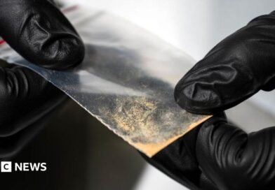Close-up of gloved hands holding a small transparent plastic packet containing a light-coloured powdery substance. The packet is being examined against an out-of-focus laboratory background.