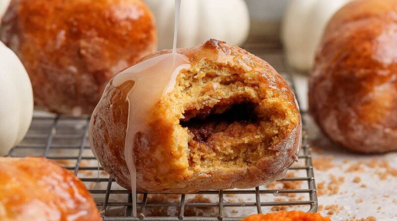 Glazed cottage cheese pumpkin fritter with a bite removed to show the spiced brown sugar center, surrounded by more fritters and small pumpkins.