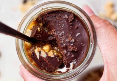 Spoon breaking through the chocolate shell on an individual Snickers cheesecake cup to reveal peanut butter and cottage cheese layers.