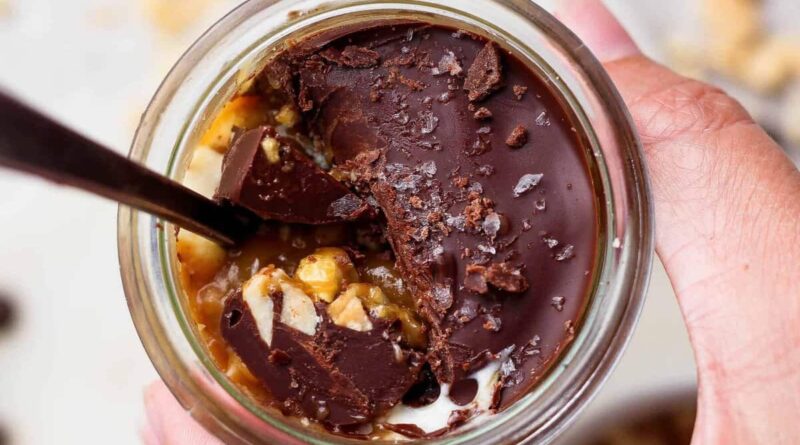 Spoon breaking through the chocolate shell on an individual Snickers cheesecake cup to reveal peanut butter and cottage cheese layers.